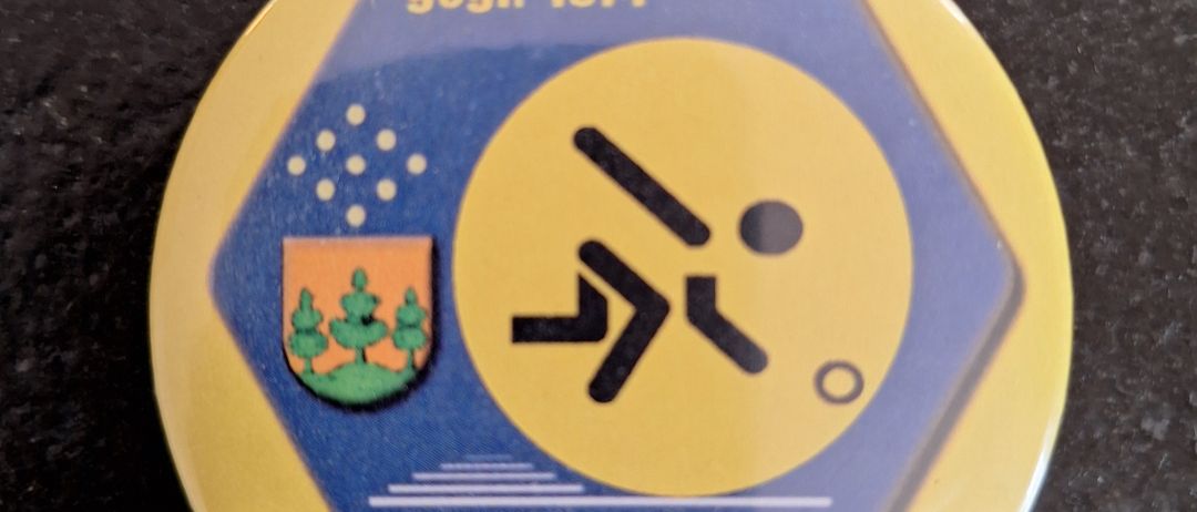 Eine gelbe und blaue kreisförmige Anstecknadel aus dem Jahr 1974, sie zeigt eine laufende Figur und ein Logo mit Bäumen, die Worte 1. KC ASKO Saalfelden stehen unten.