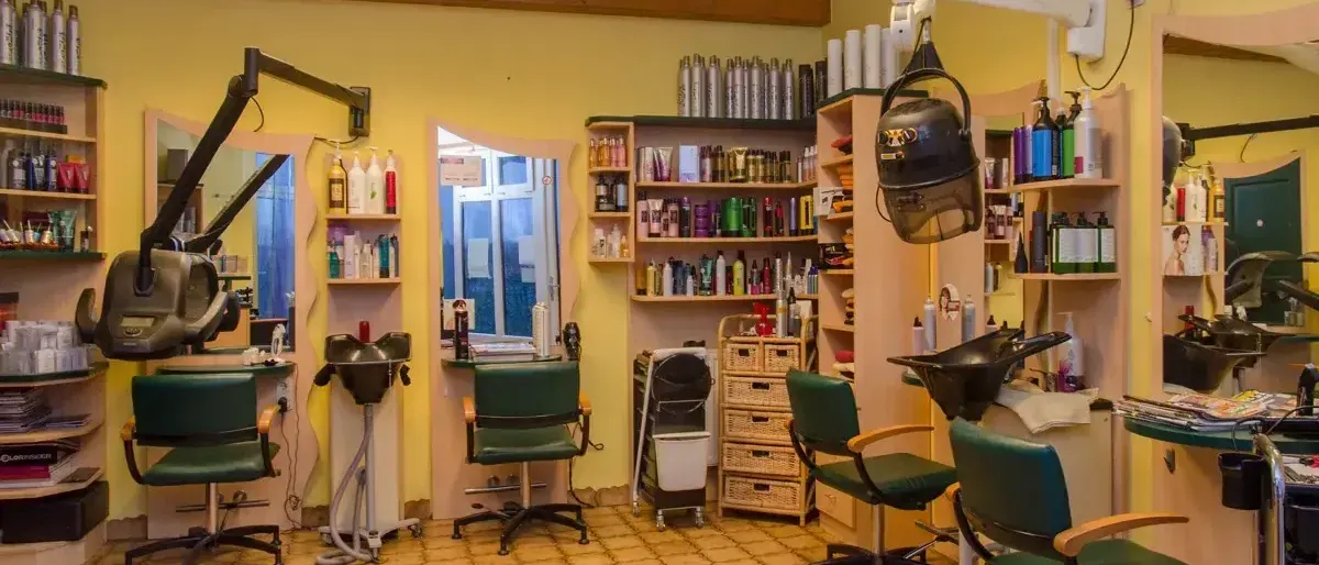 Ein Salon mit gelben Wänden hat geflieste Böden und grüne Stühle. Haartrockner und Shampooflaschen sind auf Regalen angeordnet. Ein Spiegel ist an der Wand montiert.