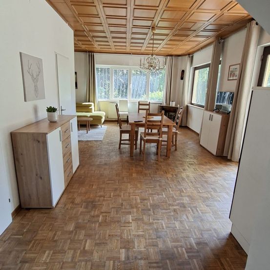 Ein gemütliches Wohnzimmer mit Holzboden, einem Esstisch und einer Kronleuchter. Ein Sofa und ein Fernseher sind im Hintergrund.