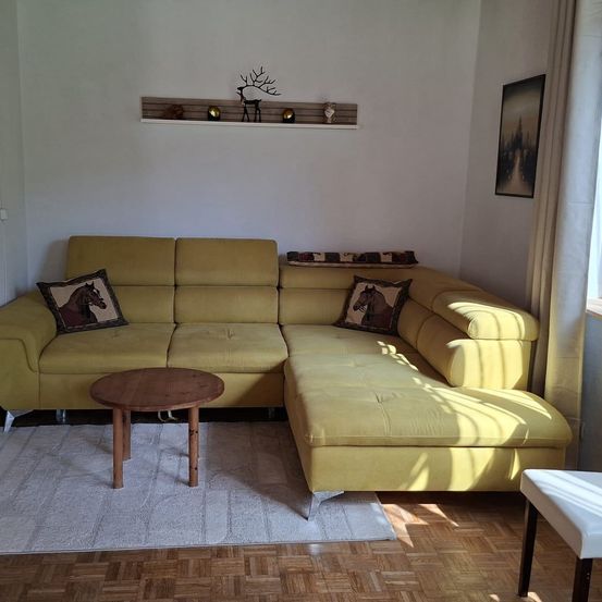 Ein Wohnzimmer mit einem gelben Ecksofa, einem runden Holztisch und einem weißen Teppich auf einem Holzboden. Ein Regal mit Dekorationsartikeln ist an der Wand über dem Sofa montiert.