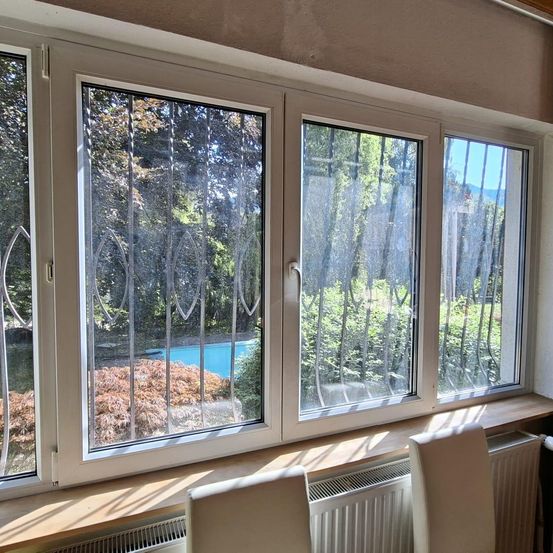 Ein Blick aus einem Zimmer mit drei großen Fenstern, die auf einen Pool und Grünflächen blicken. Die Fenster haben dekorative Metallrahmen und sind teilweise mit Jalousien verdeckt.