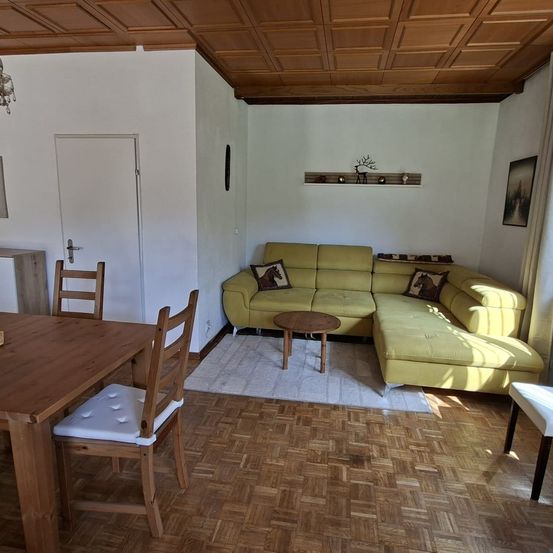 Ein Wohnzimmer mit einem gelben Sofa, einem hölzernen Esstisch und einem Holzboden. Ein weißer Teppich liegt auf dem Boden.