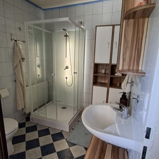 Ein Badezimmer mit einer Glasdusche, einem weißen Waschbecken und einem Holzschrank mit Regalen. Der Boden ist kariert und ein Handtuch hängt an der Wand.