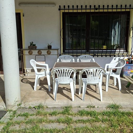 Ein Patio mit einem Holztisch und sechs weißen Stühlen für das Essen im Freien. Ein kleiner Tisch mit Topfpflanzen steht links.