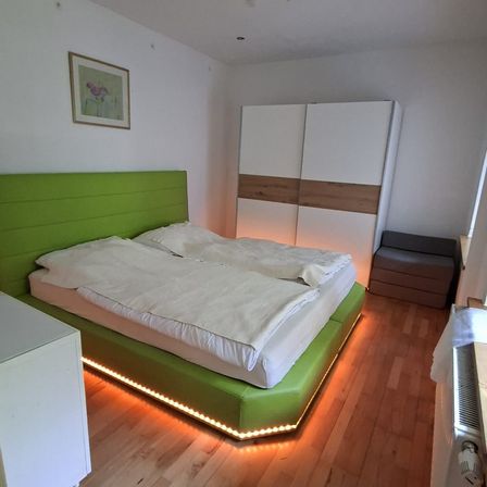 Ein modernes Schlafzimmer mit einem grünen Bettrahmen, weißen Bettwäsche und einem weißen Kleiderschrank. Der Raum hat einen Holzboden und eine weiße Wand mit einem gerahmten Bild.