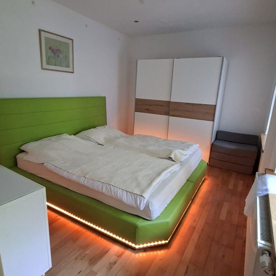 Ein modernes Schlafzimmer mit einem grünen Bettrahmen, weißen Bettwäsche und einem weißen Kleiderschrank. Der Raum hat einen Holzboden und eine weiße Wand mit einem gerahmten Bild.