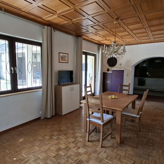 Ein gut eingerichteter Essbereich mit Holzböden, einer Kristallleuchte und einem Fernseher auf einem Schrank. Der Raum hat große Fenster und einen hölzernen Esstisch.