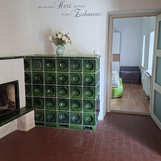 Ein gemütliches Zimmer mit Kamin, grüner Fliesenwand und einer Vase mit weißen Blumen. Im Flur befinden sich eine Couch und ein Heizkörper.