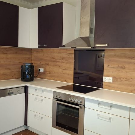 Eine moderne Küche mit Holzwänden und weißen Schränken. Ein schwarzer Induktionsherd und ein silberner Backofen stehen auf der Arbeitsplatte. Eine schwarze Kaffeemaschine ist an einer Steckdose angeschlossen.