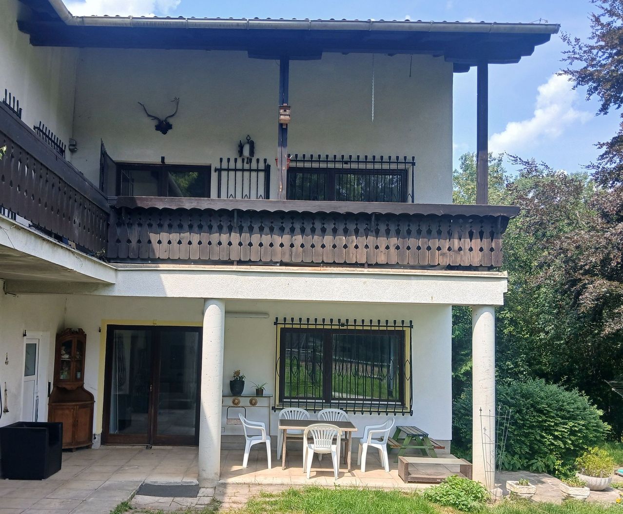 A two-story house with a balcony and a patio area with a table and chairs. The house has a deer head mounted on the wall and a potted plant on the window sill.