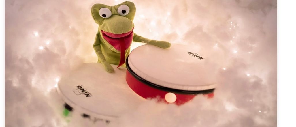 Eine Froschfigur sitzt auf zwei beleuchteten Trommeln vor einem verschneiten Hintergrund. Die Trommeln haben weiße und rote Farben und die Aufschrift 'NINO'. Ein Wasserzeichen befindet sich unten rechts.