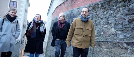 Drei Personen, zwei Männer und eine Frau, gehen eine Kopfsteinpflasterstraße entlang. Sie sind für kaltes Wetter warm gekleidet, mit Schals und Mänteln. Der Mann auf der rechten Seite lächelt und schaut direkt in die Kamera. Hinter ihnen befindet sich eine Steinmauer mit einer Backsteinstruktur.