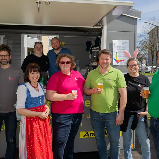 Eine Gruppe von Menschen steht vor einem Foodtruck, lächelt und hält Getränke in den Händen. Eine Frau trägt ein traditionelles bayerisches Kleid. Dahinter wirbt ein Schild für ein Festival.