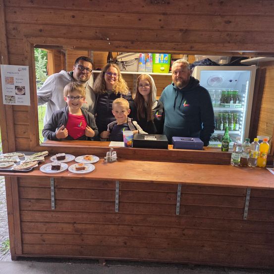 Eine Familie mit fünf Personen steht lächelnd hinter einem Tresen. Sie befinden sich in einem Holzkiosk mit einem Kühlschrank voller Getränke. Vor ihnen stehen Teller mit Essen, einschließlich Kuchen. An der Wand befindet sich ein Schild mit Preisen.