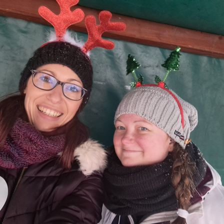 Zwei Frauen lächeln in Winterkleidung, eine mit roten Rentierhörnern und die andere mit Weihnachtsbäumen auf ihrer Mütze. Beide sind drinnen.
