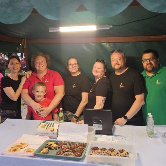 Eine Gruppe von Menschen steht hinter einem Tisch unter einem Zelt mit verschiedenen Desserts, darunter Kekse und Cupcakes. Sie tragen passende Kleidung, und ein Kind sitzt in der Mitte. Ein Laptop und eine Wasserflasche sind auf dem Tisch.