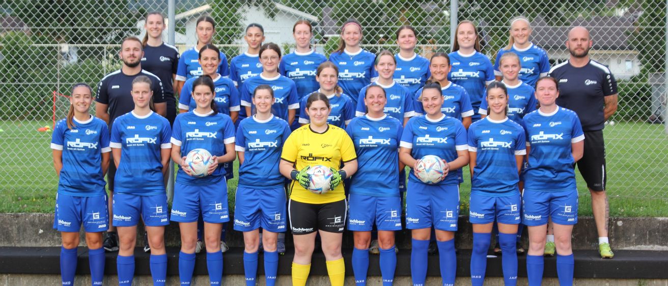 Eine Frauenfußballmannschaft, gekleidet in blauen Trikots mit 'Redfix' auf ihren Jerseys, posiert für ein Gruppenfoto. Eine Spielerin in Gelb hält zwei Fußballbälle, während andere einen halten. Sie stehen auf einem Feld mit einem Zaun im Hintergrund.
