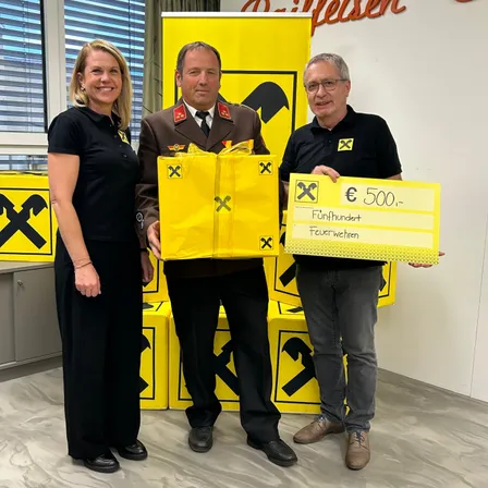 Drei Personen stehen vor einem gelben Hintergrund und halten eine gelbe Tasche und ein Schild mit 500 €. Der Mann in der Mitte trägt einen Militäruniform, während die anderen schwarze Shirts tragen.