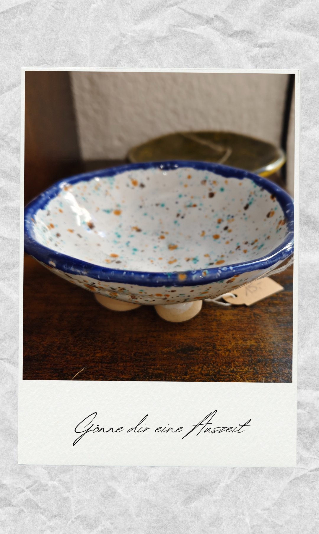 Eine farbenfrohe, gefleckte Schüssel mit blauem Rand steht auf einem Holztisch mit einem hängenden Etikett.