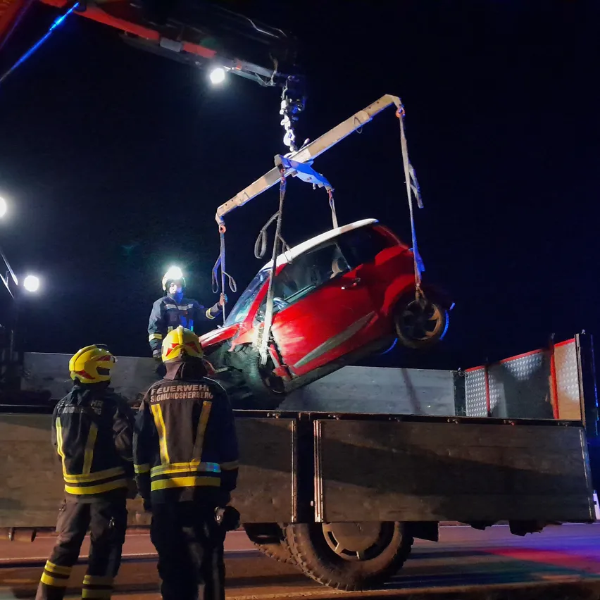 Drei Feuerwehrleute in gelben Helmen heben nachts ein rotes Auto mit einem Kran. Das Auto ist auf dem Kopf und das Vorderrad ist abgekoppelt.