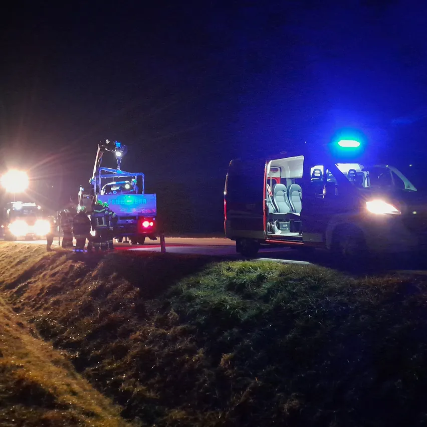 Notfallfahrzeuge stehen nachts am Straßenrand. Die Lichter sind eingeschaltet, und eine Person steht in der Nähe der Fahrzeuge.