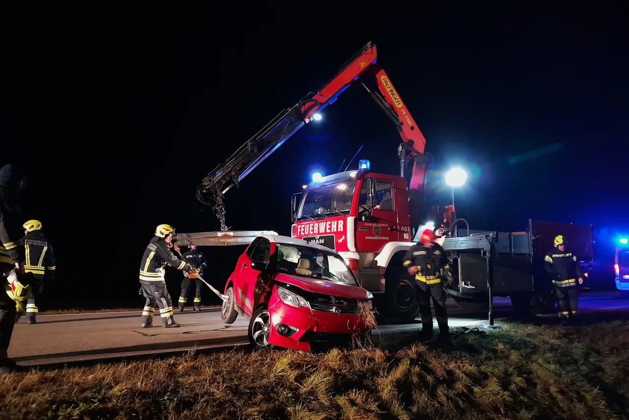 Feuerwehrleute verwenden einen Kran, um ein beschädigtes rotes Auto von der Straße zu heben. Die Szene spielt bei Nacht, wobei zwei uniformierte Personen die Operation durchführen.