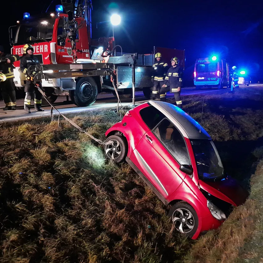 Ein rotes Auto ist nachts in einen Grasbereich gekracht. Rettungsdienste sind vor Ort mit einem Feuerwehrwagen und einem Krankenwagen.