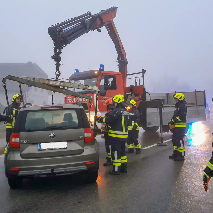 Ein roter Kran hebt ein Auto an, das von Feuerwehrleuten in gelben Uniformen umgeben ist, auf einer nebligen Straße.