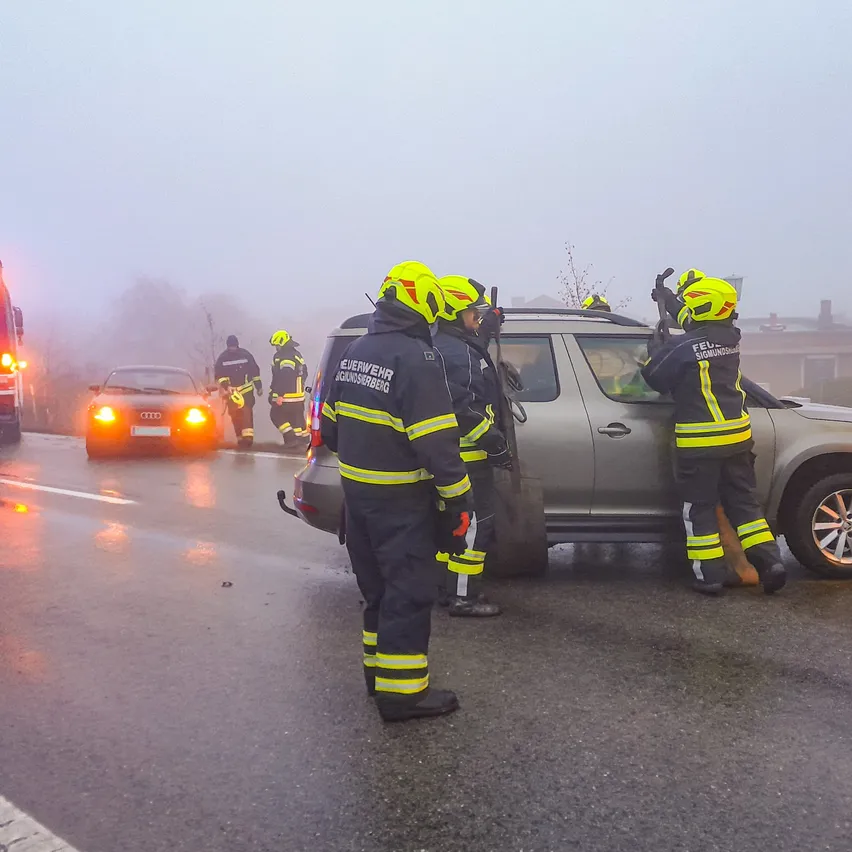 Feuerwehrleute helfen bei einem Unfall an der Straße. Sie tragen gelbe und schwarze Uniformen. Einer hält die Autotür, während ein anderer in der Nähe des Fahrzeugs steht. Die Szene ist neblig.