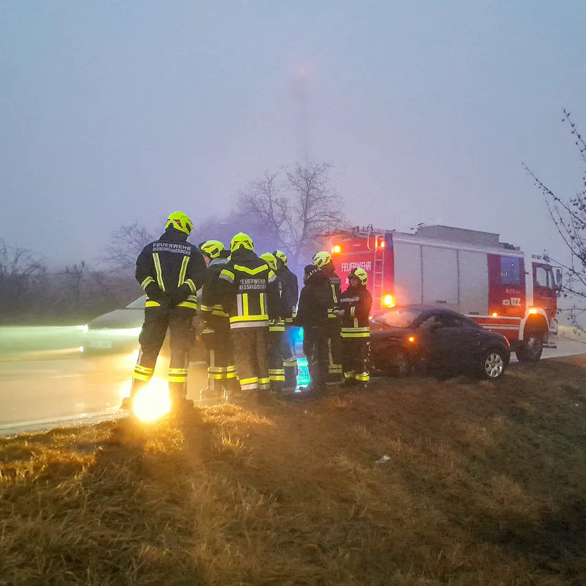 Einsatzkräfte stehen am Straßenrand, beleuchtet von einer Taschenlampe, in der Nähe eines beschädigten Autos und eines Feuerwehrwagens mit eingeschalteten Lichtern.