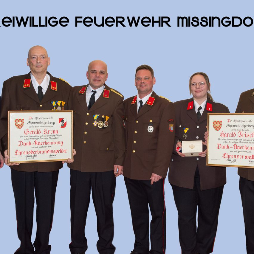 Fünf Personen in Militäruniformen stehen zusammen und halten Urkunden. Die beiden links und rechts halten größere gerahmte Urkunden. Im Hintergrund steht 'Eiwillige Feuerwehr Missingdon'.