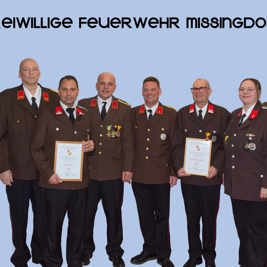 Sechs Personen in Uniform posieren für ein Foto. Zwei Männer in der Mitte halten Urkunden, während die anderen hinter ihnen stehen. Der Hintergrund ist blau mit weißem Text, der 'Freiwillige Feuerwehr Missingdorf' lautet.