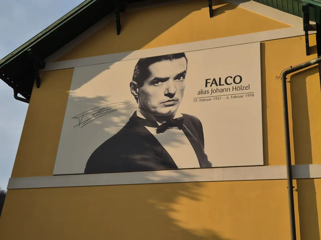 Ein Poster von Falco, auch bekannt als Johann Holzel, ist an der Seite eines gelben Gebäudes. Das Poster enthält eine Unterschrift.