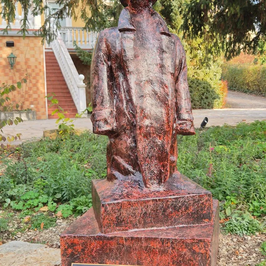Eine Bronzestatue eines Mannes in einem Mantel steht auf einem roten Marmorpodest in einem Garten. Die Statue befindet sich vor einem Haus mit einer Treppe.