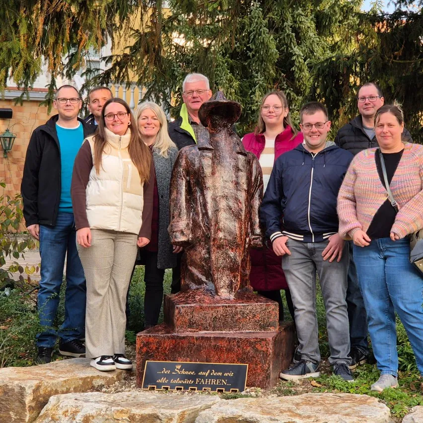 Eine Gruppe von Menschen steht vor einer Statue mit einer Plakette auf einem Steinsockel und lächelt für ein Foto. Die Statue stellt einen Mann in einem Hut dar. Dahinter befinden sich Bäume und ein Gebäude.