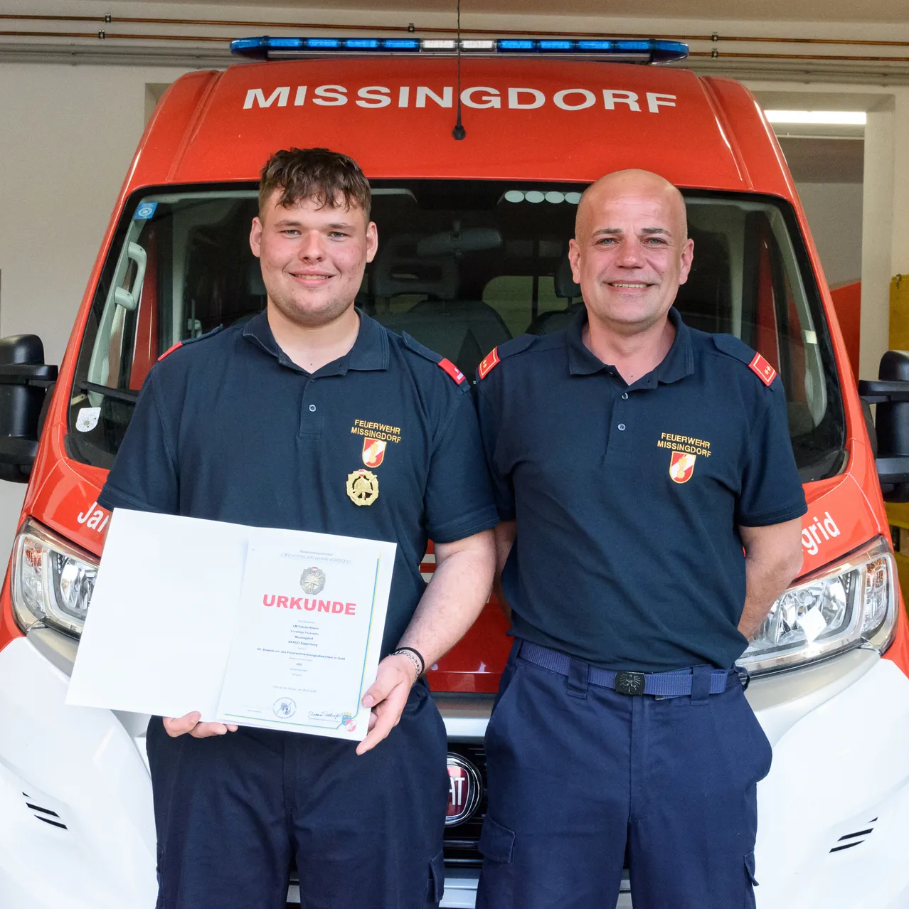 Zwei Feuerwehrleute in Uniform stehen vor einem roten Feuerwehrwagen, einer hält eine Urkunde in der Hand. Der Mann auf der linken Seite lächelt und der Mann auf der rechten Seite schaut in die Kamera.