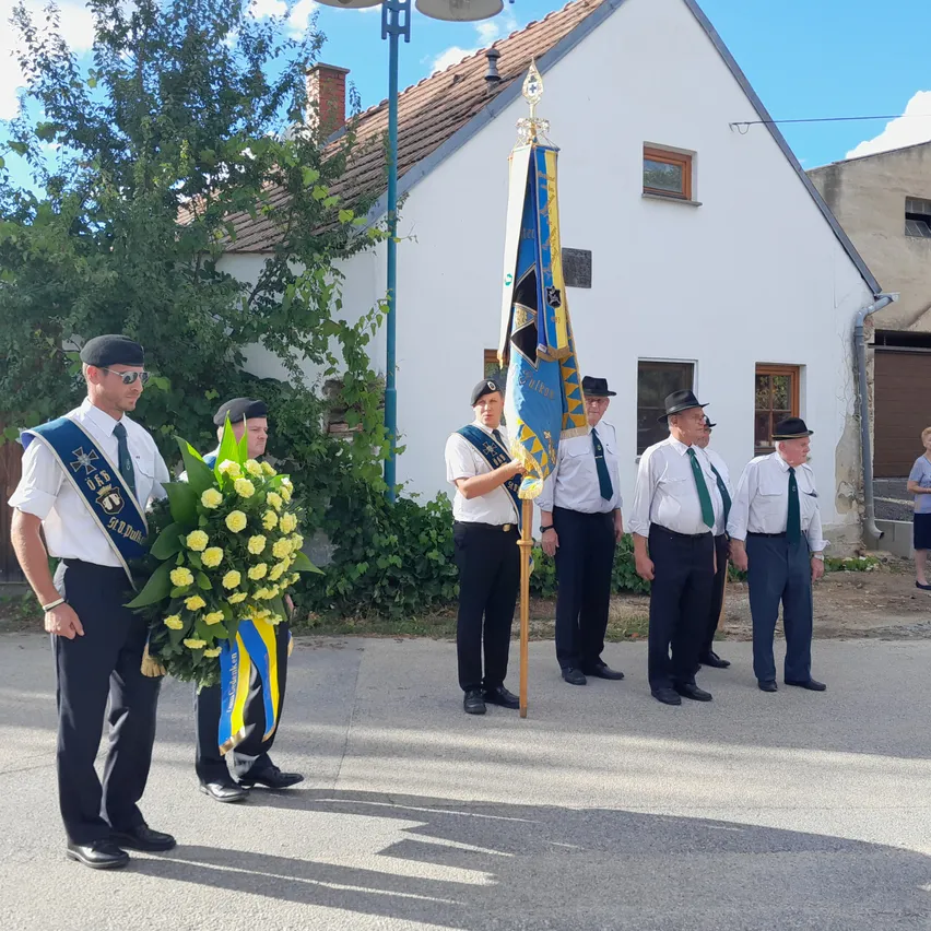 Eine Gruppe von Männern in Uniformen hält eine Flagge und einen Kranz und steht an einem sonnigen Tag vor einem Haus.
