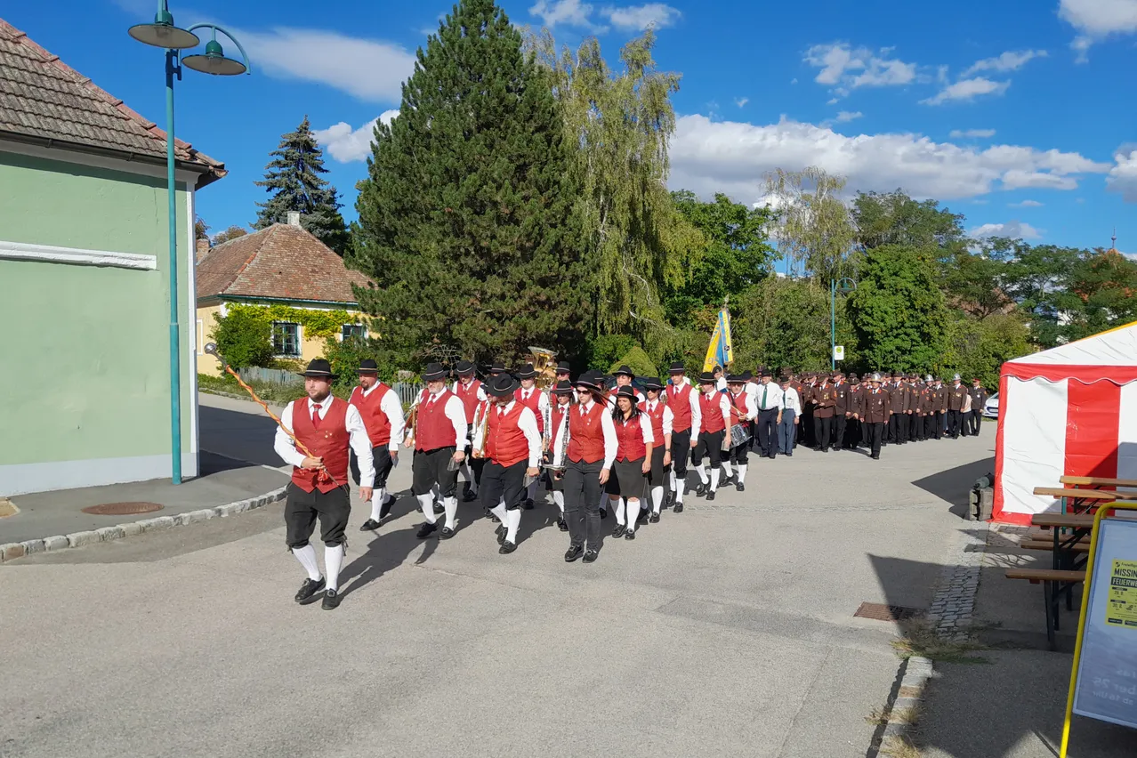 Eine Blaskapelle in traditioneller Kleidung marschiert die Straße entlang, begleitet von einer Fahne und umgeben von Zuschauern.