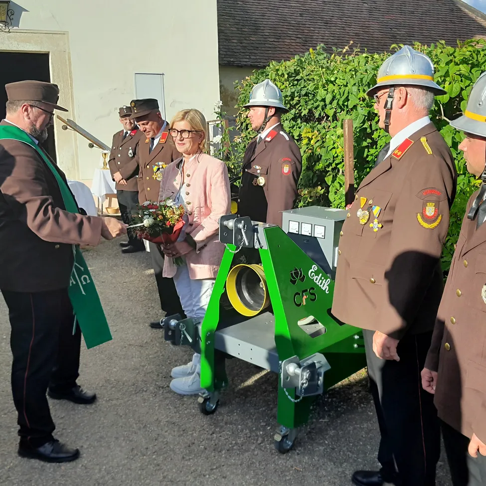 Eine Gruppe uniformierter Personen, einschließlich einer Frau in einer rosa Jacke, stehen um eine grüne Maschine namens Edith CFS. Sie sind vor einem Gebäude, wo ein Mann der Frau einen Blumenstrauß überreicht.