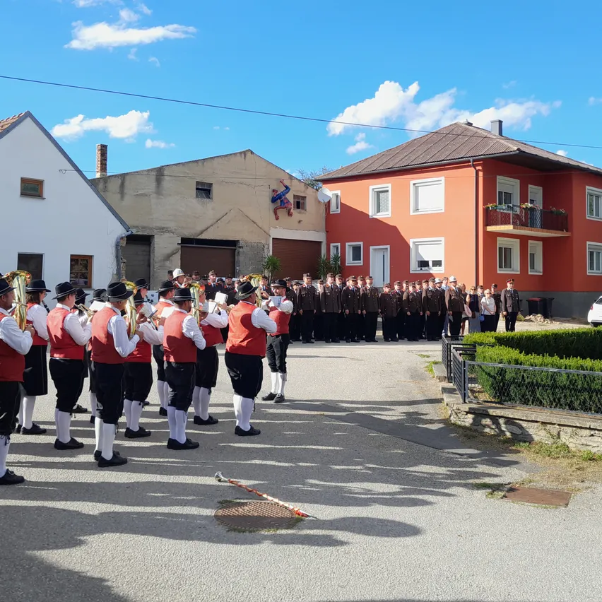 Eine Gruppe von Musikern in traditioneller Kleidung steht vor einem Gebäude an einem sonnigen Tag. Dahinter ist eine Reihe von Menschen in Uniformen zu sehen.