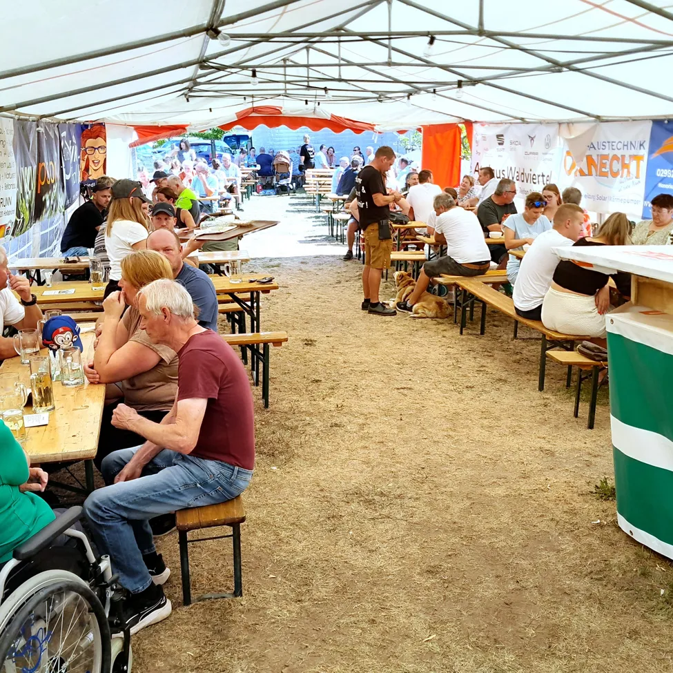 Eine Gruppe von Menschen sitzt auf Bänken unter einem Zelt, einige trinken und andere unterhalten sich. Ein Mann im Rollstuhl sitzt vor einem Tisch.
