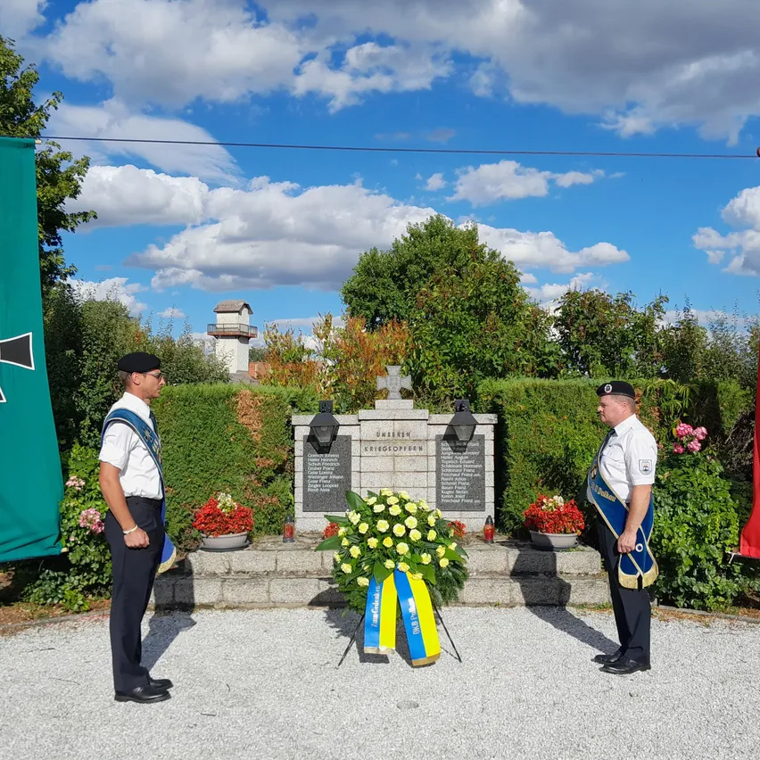 Zwei Männer in Uniform stehen vor einem Denkmal mit einem Kreuz und einer Girlande, umgeben von Blumen und Grün.
