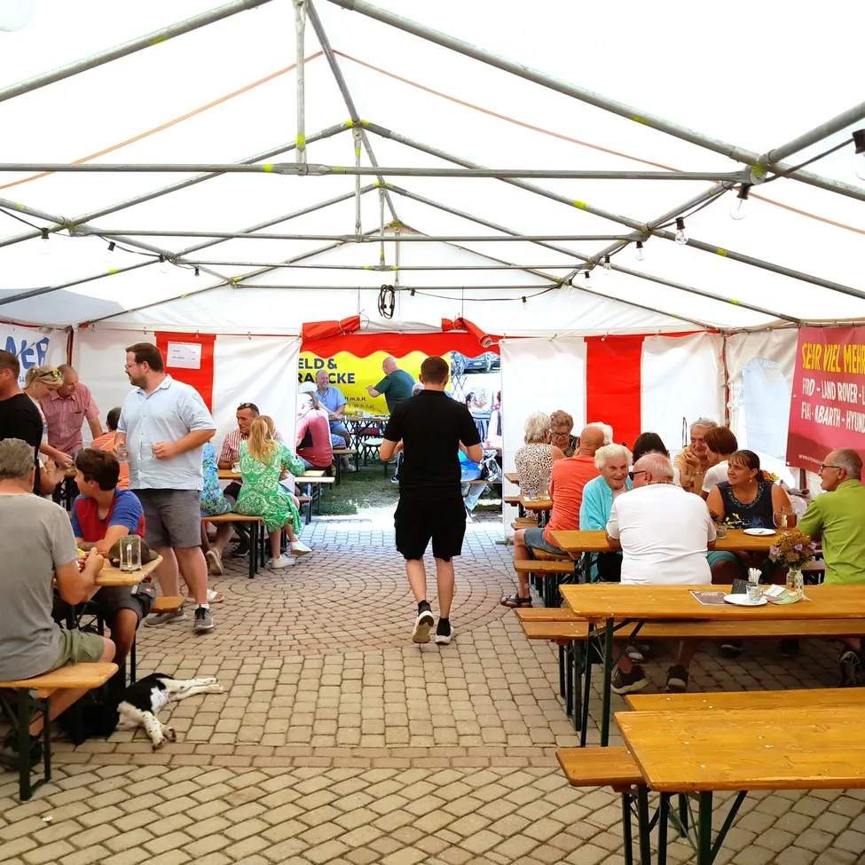 Ein großes Zelt mit Tischen und Bänken für ein Outdoor-Essevent. Die Leute sitzen, essen und unterhalten sich. Ein Mann geht durch die Mitte, und ein Hund liegt auf dem Boden. Banner sind an den Seiten des Zeltes angebracht.