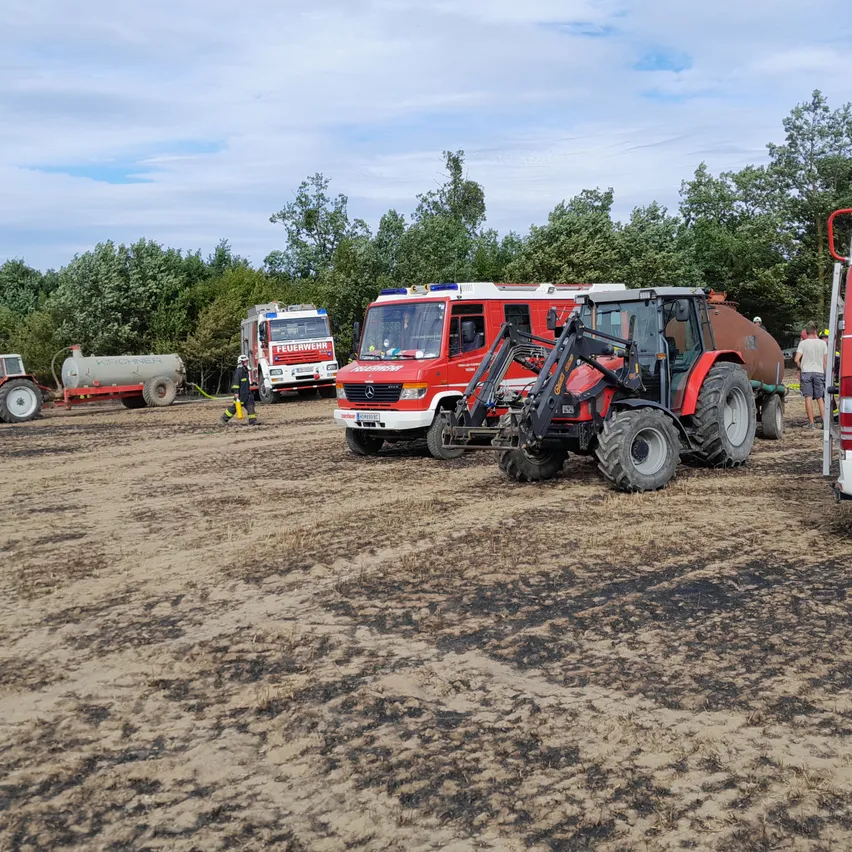Mehrere Fahrzeuge, darunter Feuerwehrwagen und Traktoren, sind auf einem Feld mit Erdspuren und Brandflecken geparkt.