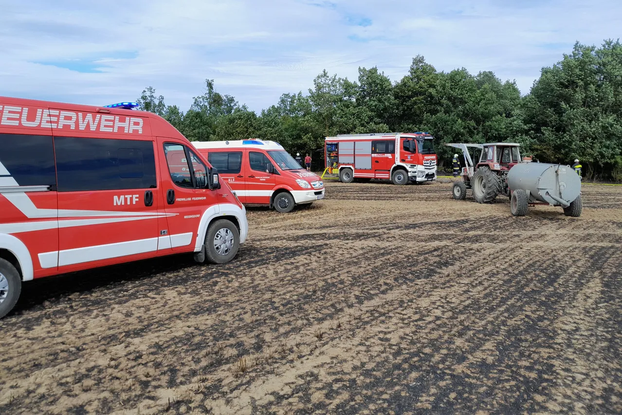 Rettungsfahrzeuge stehen auf einem schlammigen Feld, mit Bäumen und einem Traktor im Hintergrund. Eine Person steht neben einem der Fahrzeuge.