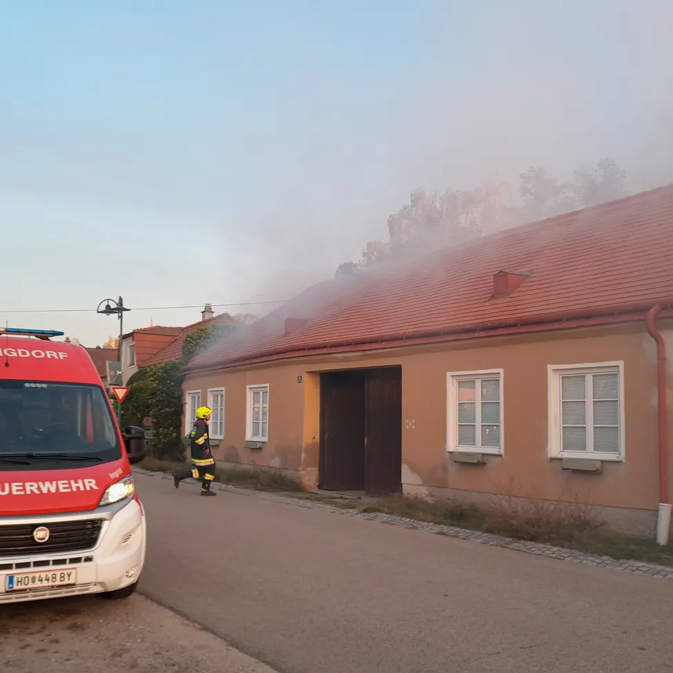 Ein roter und weißer Feuerwehrwagen ist am Straßenrand geparkt. Ein Feuerwehrmann in voller Ausrüstung rennt auf ein Haus zu, aus dem Rauch aufsteigt.
