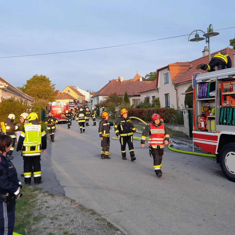 Eine Gruppe von Feuerwehrleuten ist auf einer Straße versammelt, einige tragen Helme und tragen Ausrüstung. Ein Feuerwehrwagen ist am Straßenrand geparkt, mit Feuerwehrleuten oben.