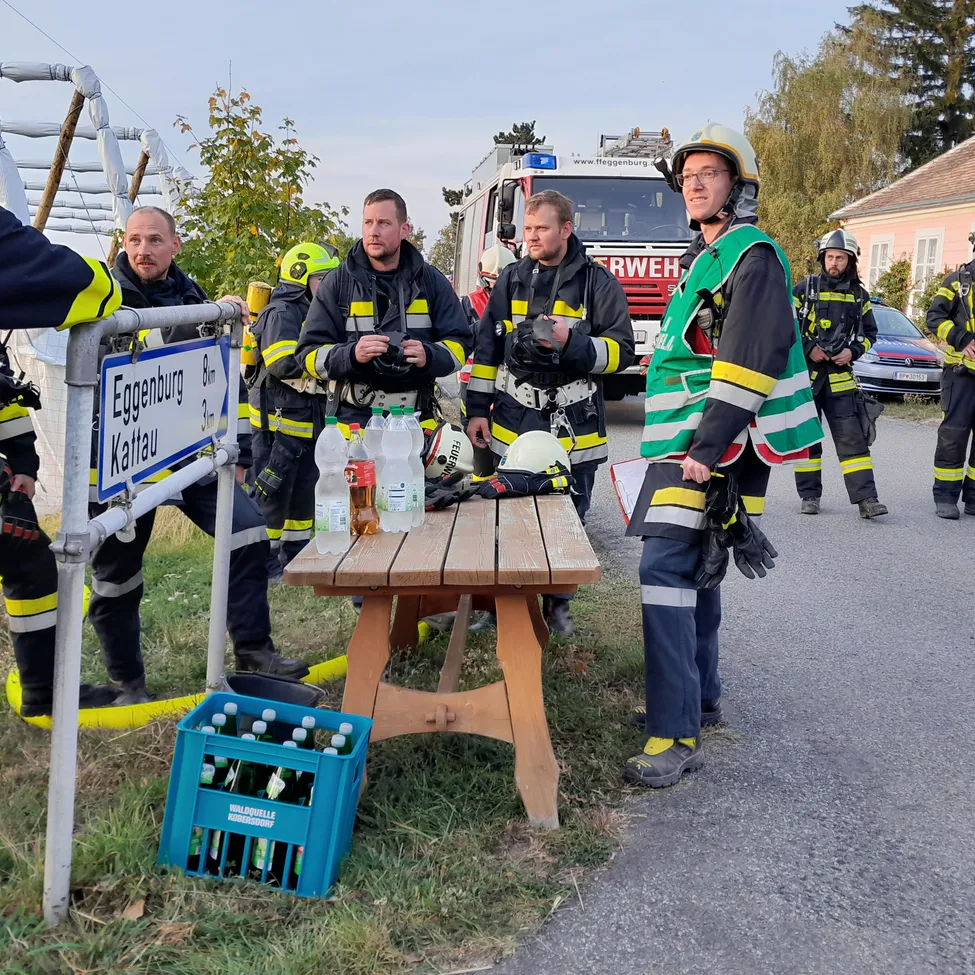 Eine Gruppe von Feuerwehrleuten steht am Straßenrand, einige tragen Helme und andere stehen um einen Tisch mit Flaschen und einem Kasten Bier.