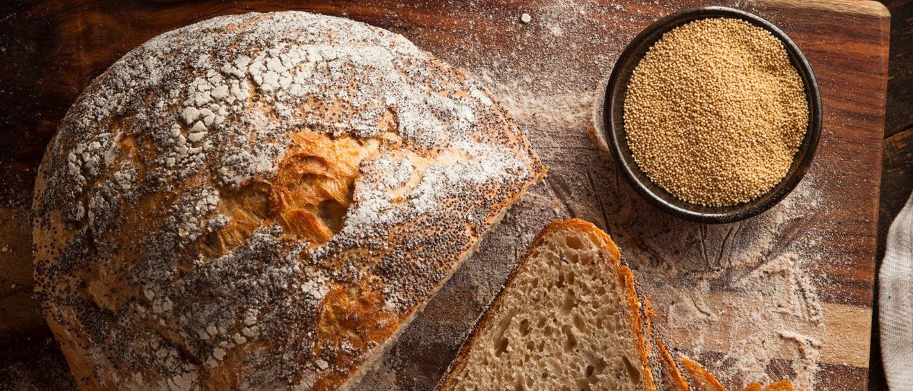 Ein Laib rustikales Brot, geschnitten auf einem Holzschneidebrett, mit einer Schüssel Samen in der Nähe. Im Hintergrund befinden sich Mehlschüsseln und ein Handtuch.