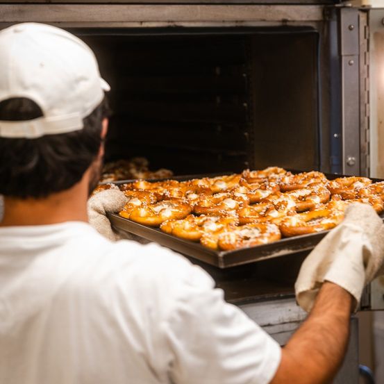 Eine Person in einer weißen Mütze und Handschuhen nimmt eine Schale mit Brezeln aus einem Ofen.